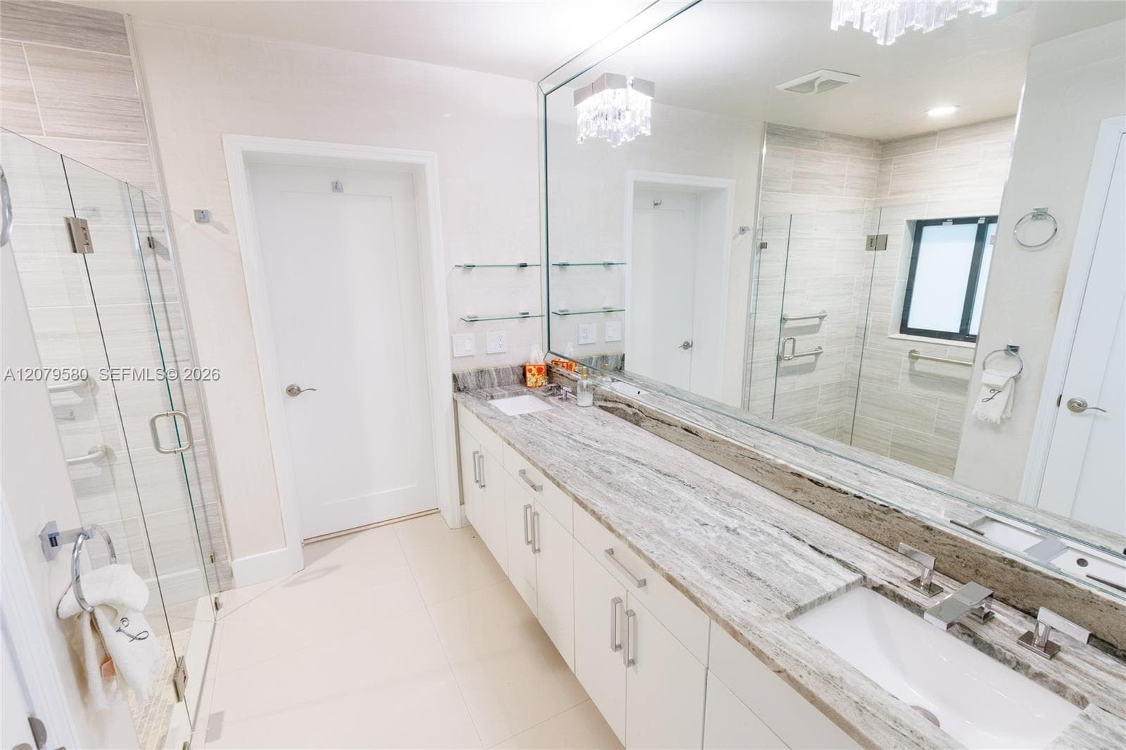 Double vanity with natural stone countertops, dual under-mount sinks, expansive wall-to-wall mirror, glass-enclosed shower with horizontal tile surround, and tile flooring