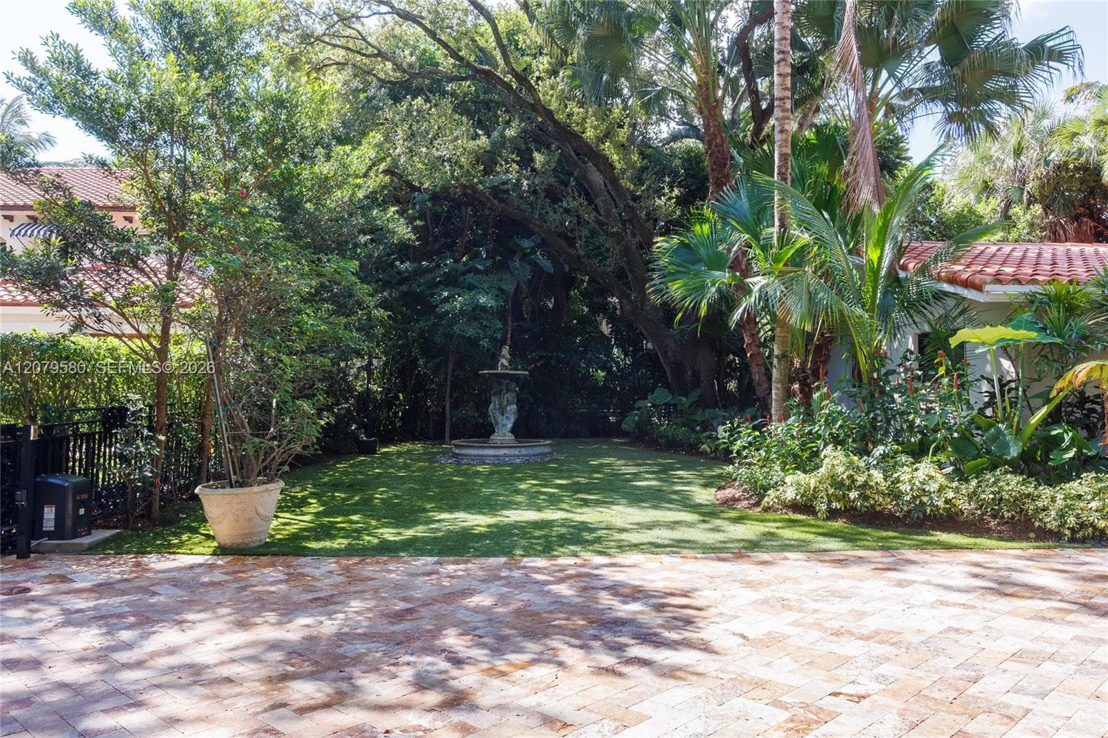 Landscaped grounds featuring a stone-paver driveway, manicured lawn, tiered water fountain, and mature tropical foliage