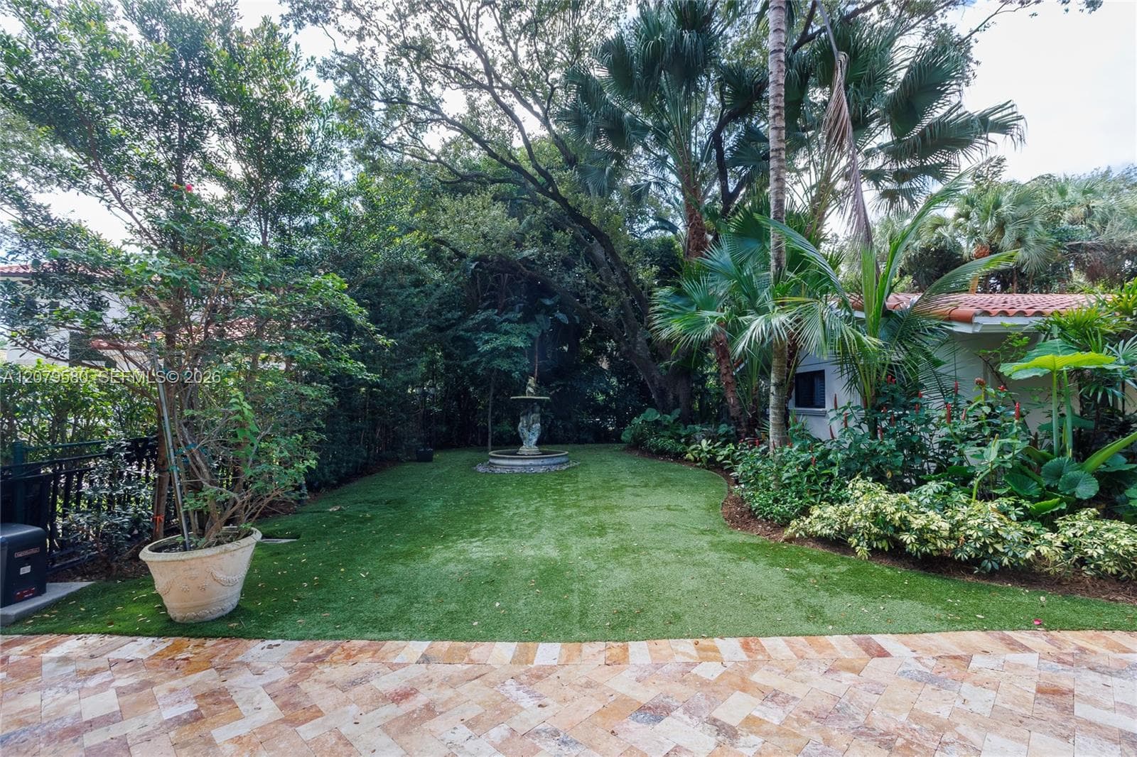 Backyard featuring a paved stone patio in a herringbone pattern, a central tiered fountain, mature trees, manicured lawn, and established tropical landscaping