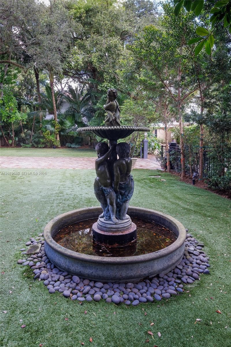 Tiered sculptural stone fountain featuring a basin surrounded by river rock landscaping, set within a grass lawn under a canopy of mature trees