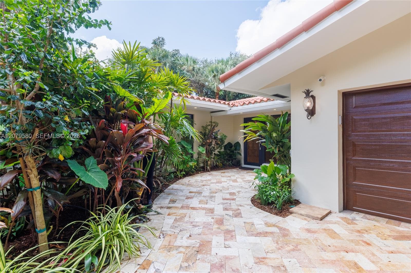 Stucco exterior featuring a herringbone-pattern stone paver walkway, clay tile roof, wood-finish garage door, and recessed exterior lighting