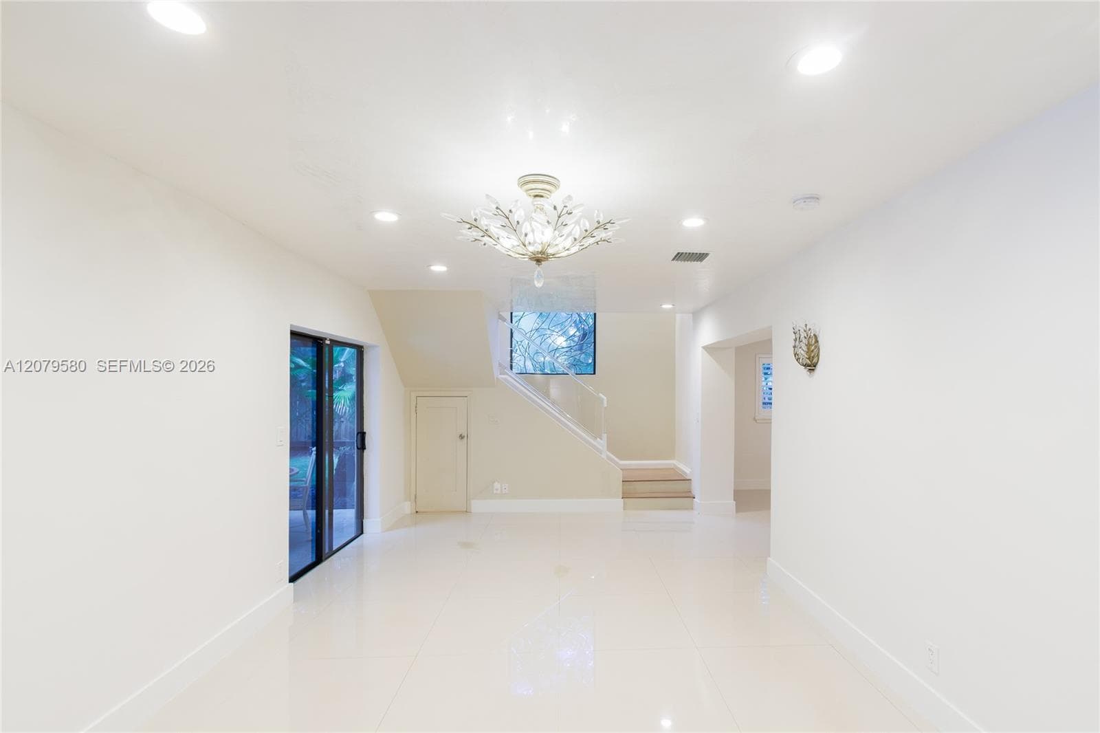 Open-concept interior featuring large-format tile flooring, recessed lighting, a decorative crystal-style chandelier, and a staircase with a glass panel railing