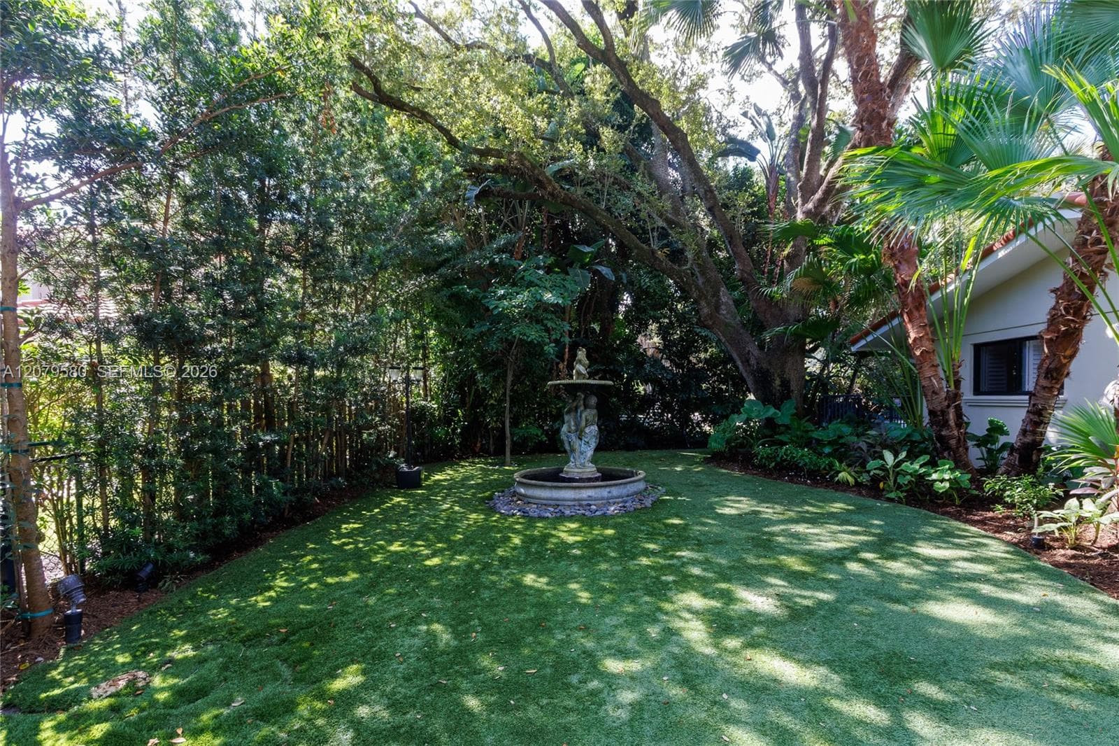 Backyard featuring a tiered stone fountain, mature trees, dense perimeter landscaping, and a manicured lawn