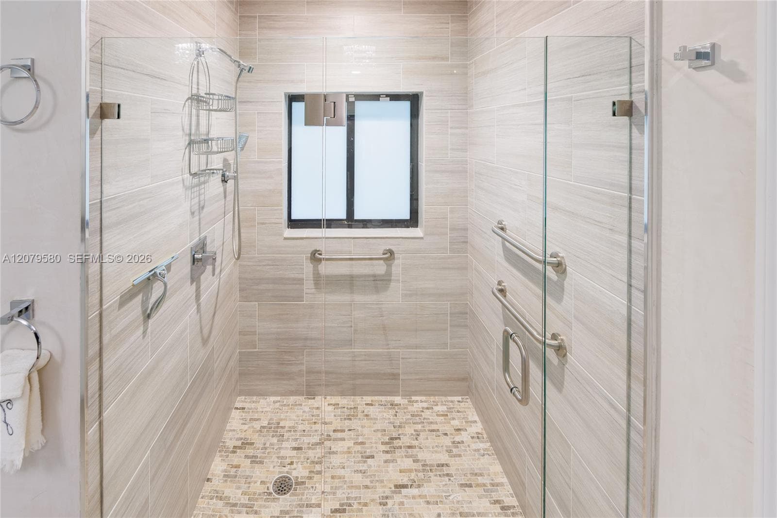 Walk-in shower featuring floor-to-ceiling neutral tile, mosaic stone-finish flooring, glass panel enclosure, and stainless steel hardware