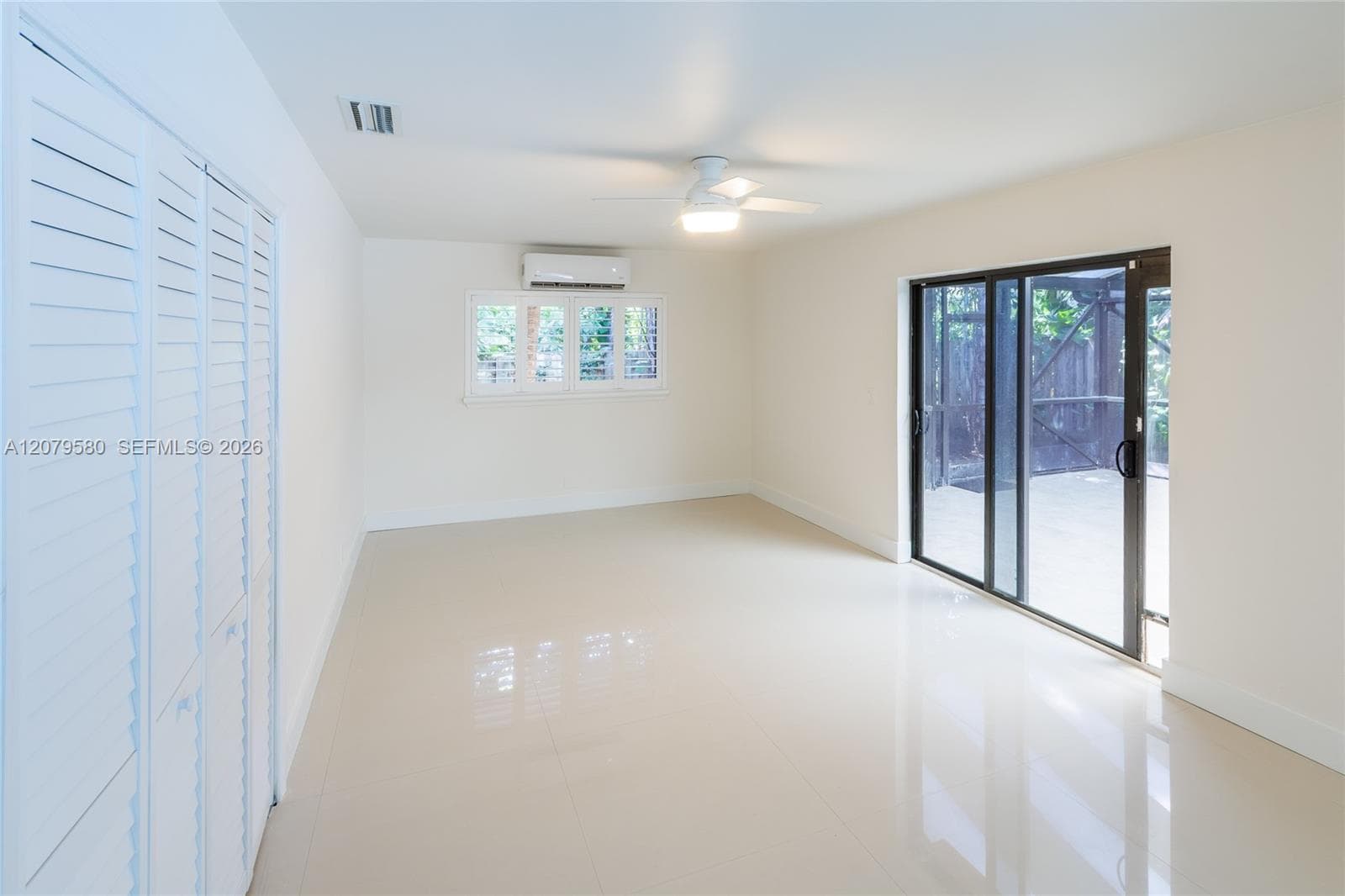 Neutral-toned tile flooring, white louvered-door closets, wall-mounted climate control unit, ceiling fan with integrated lighting, and glass sliding doors