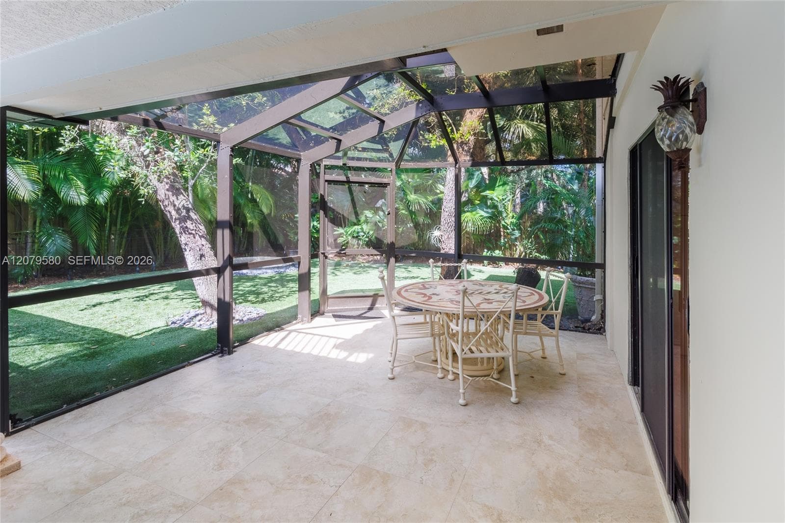 Screened-in lanai featuring large-format stone tile flooring, a geometric metal frame enclosure, wall-mounted exterior decorative lighting, and a lawn with mature tropical landscaping