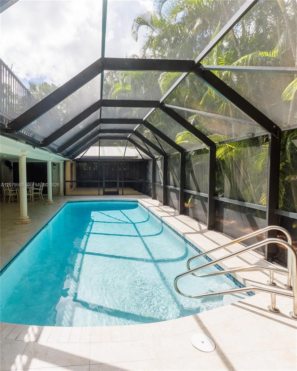 Screen-enclosed swimming pool featuring a light-toned paved deck, metal handrails, and structural pillars