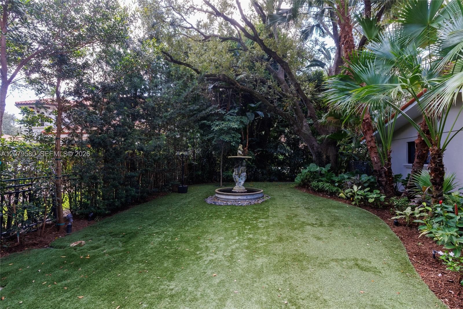 Landscaped backyard featuring a central tiered stone fountain, mature perimeter trees, lush palms, and a metal perimeter fence