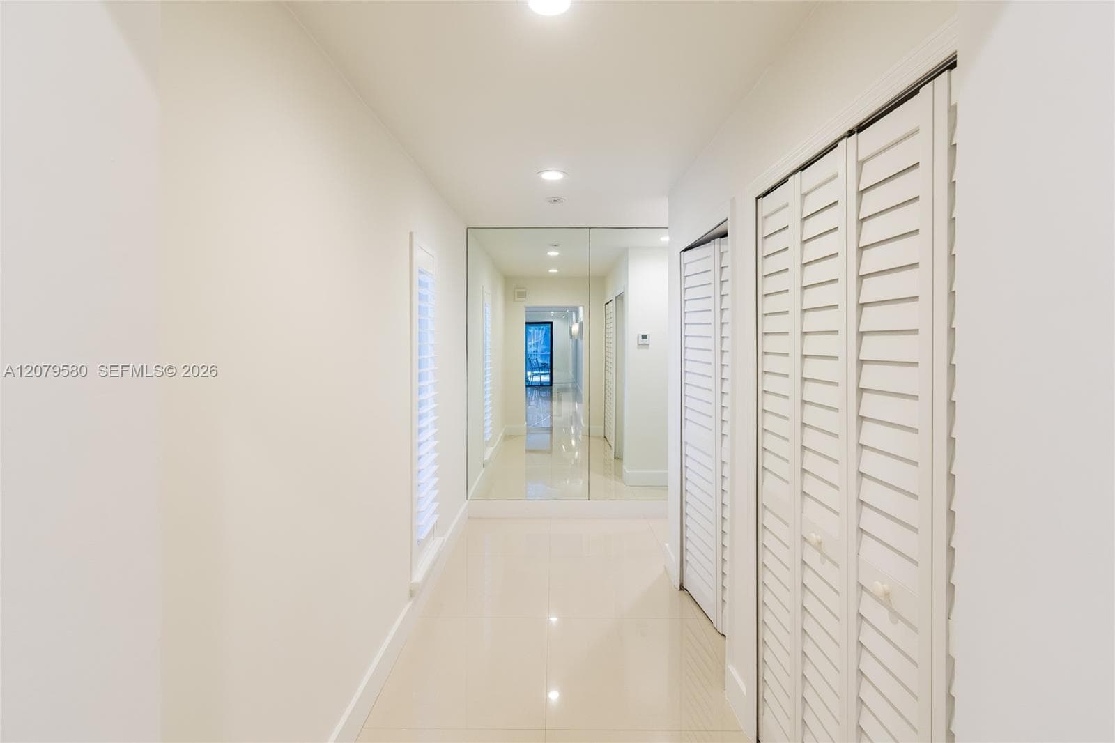 Hallway featuring neutral polished tile flooring, floor-to-ceiling mirror paneling, recessed lighting, and white louvered closet doors