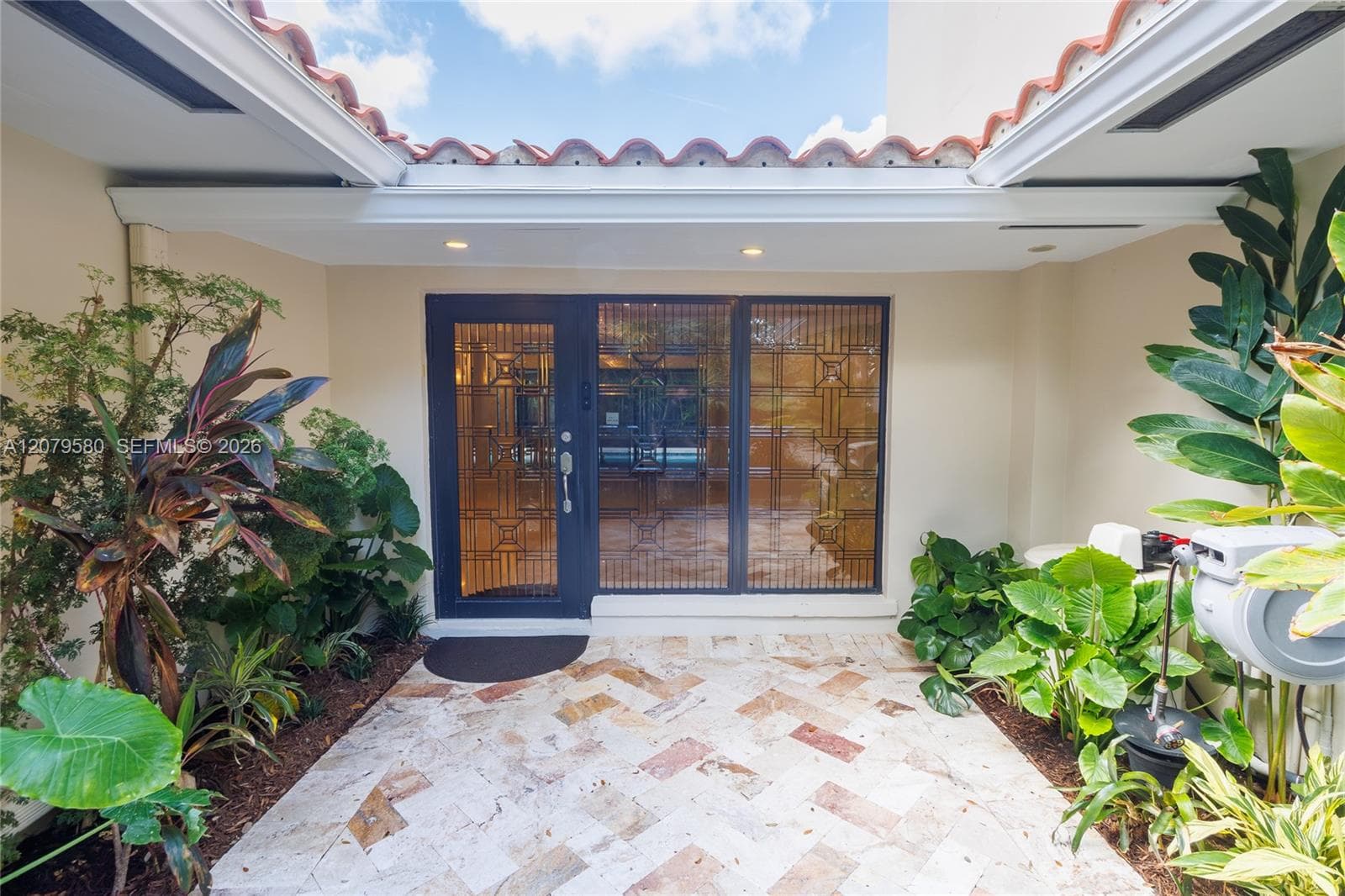 Courtyard entrance featuring herringbone-patterned stone flooring, a glass-paneled front door with geometric metal detailing, recessed lighting, and a red tile roofline