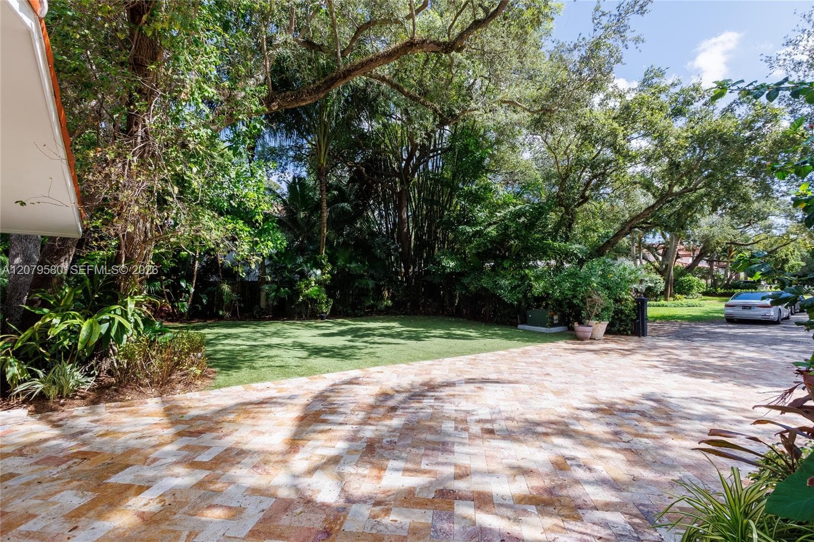Expansive paved driveway featuring variegated stone-finish tiles, synthetic grass lawn, and established mature trees