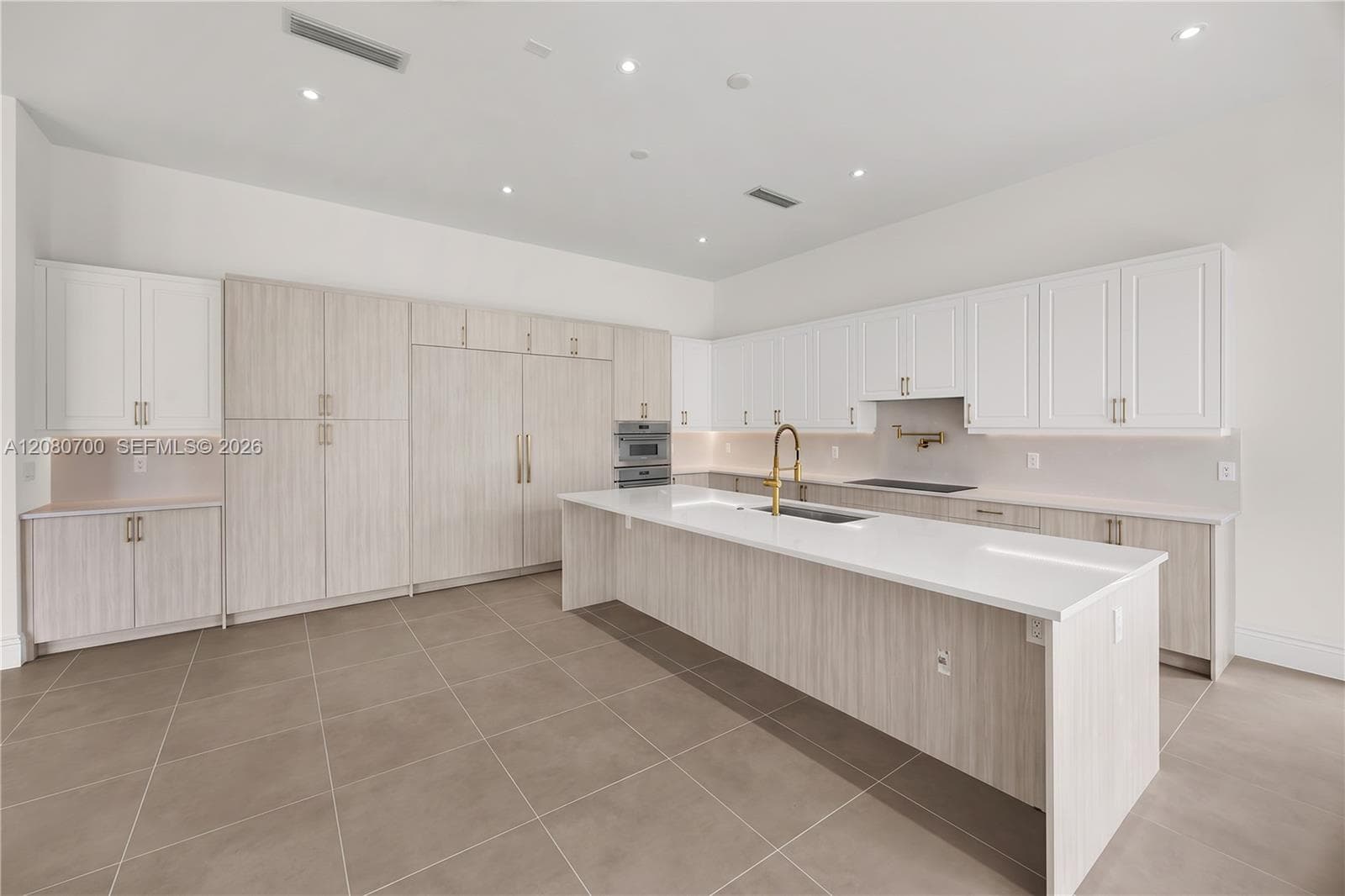 Expansive kitchen featuring a large central island with a sink, extensive white cabinetry, light wood-finish cabinetry, built-in stainless steel appliances, and sleek tile flooring