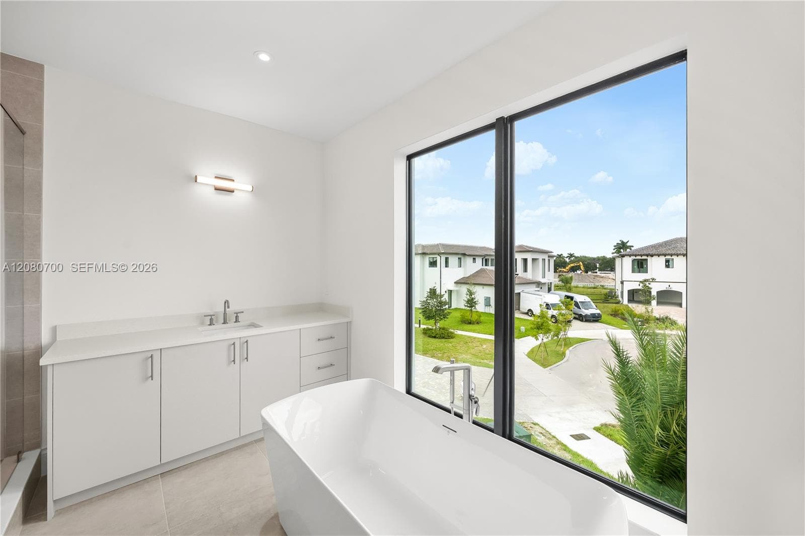 Freestanding soaking tub, large window, vanity with light-colored countertop, recessed lighting, and tile flooring