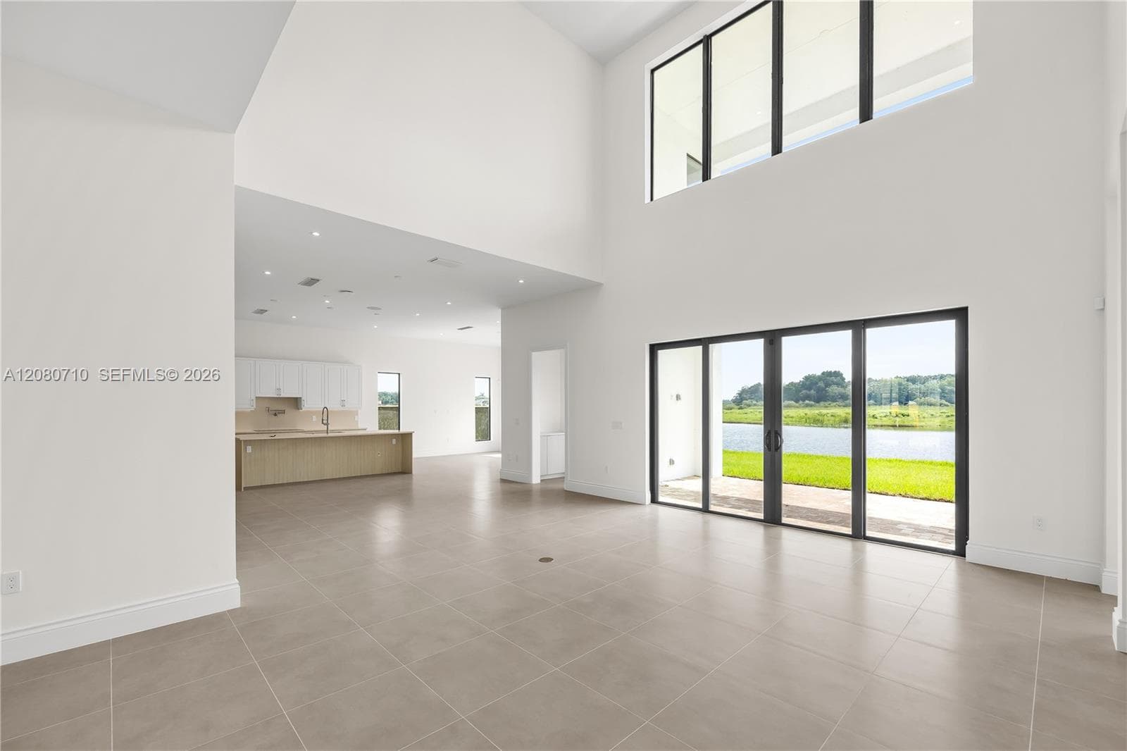 Expansive open-concept living space featuring soaring ceilings, abundant natural light from floor-to-ceiling glass sliding doors and upper clerestory windows, polished concrete flooring throughout, and a seamlessly integrated kitchen with wood-finish cabinetry visible in the background