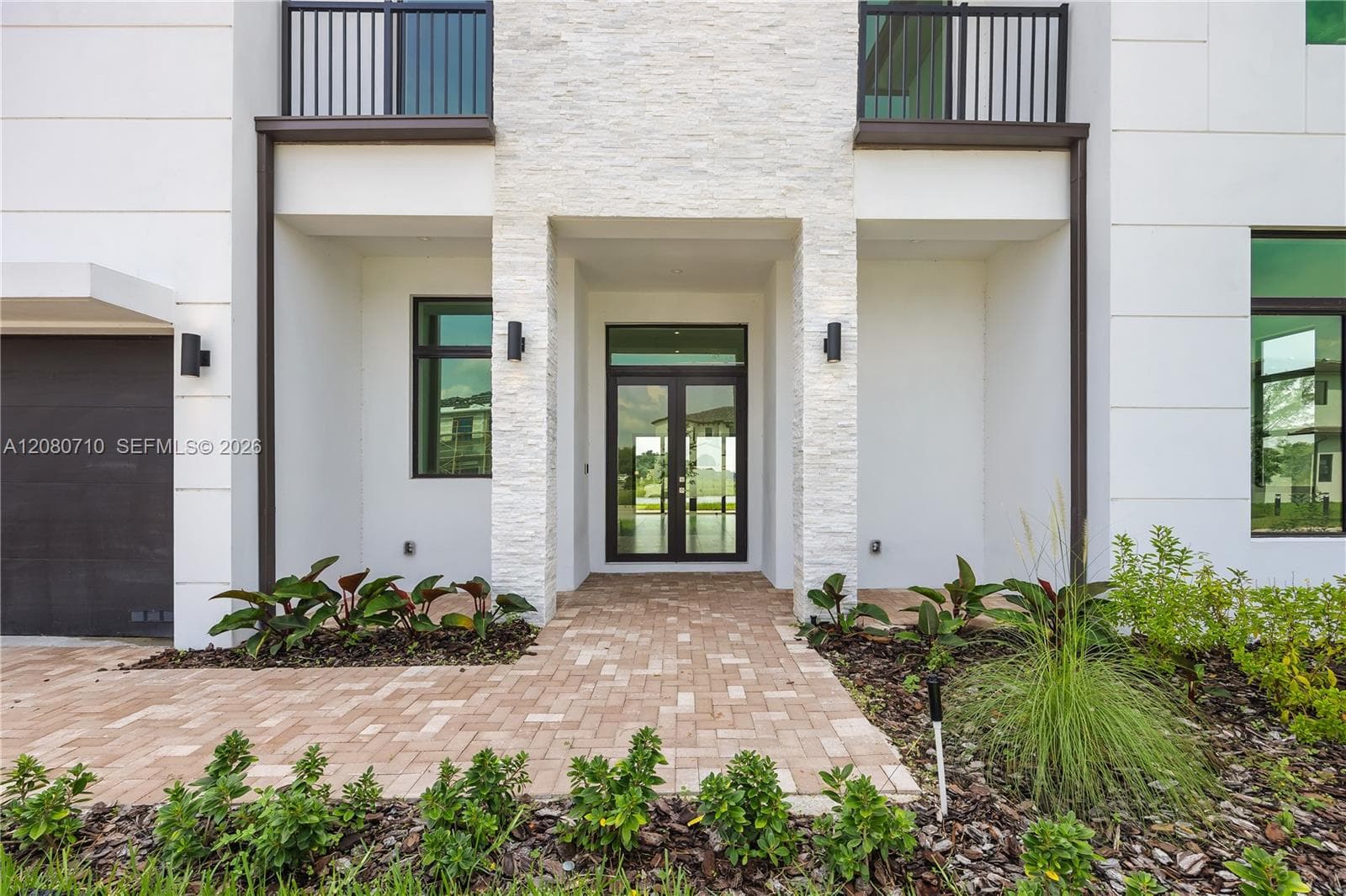 Modern exterior with stacked stone accents, double entry doors, dark frame windows, integrated exterior lighting, and a paver walkway