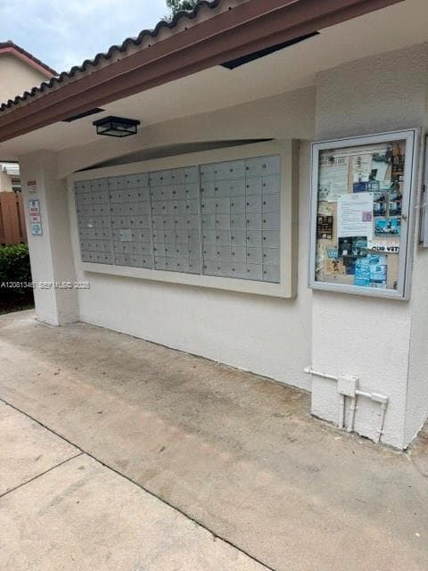 Mailbox station featuring multiple lockable compartments, exterior lighting, a tiled roof overhang, and a community bulletin board