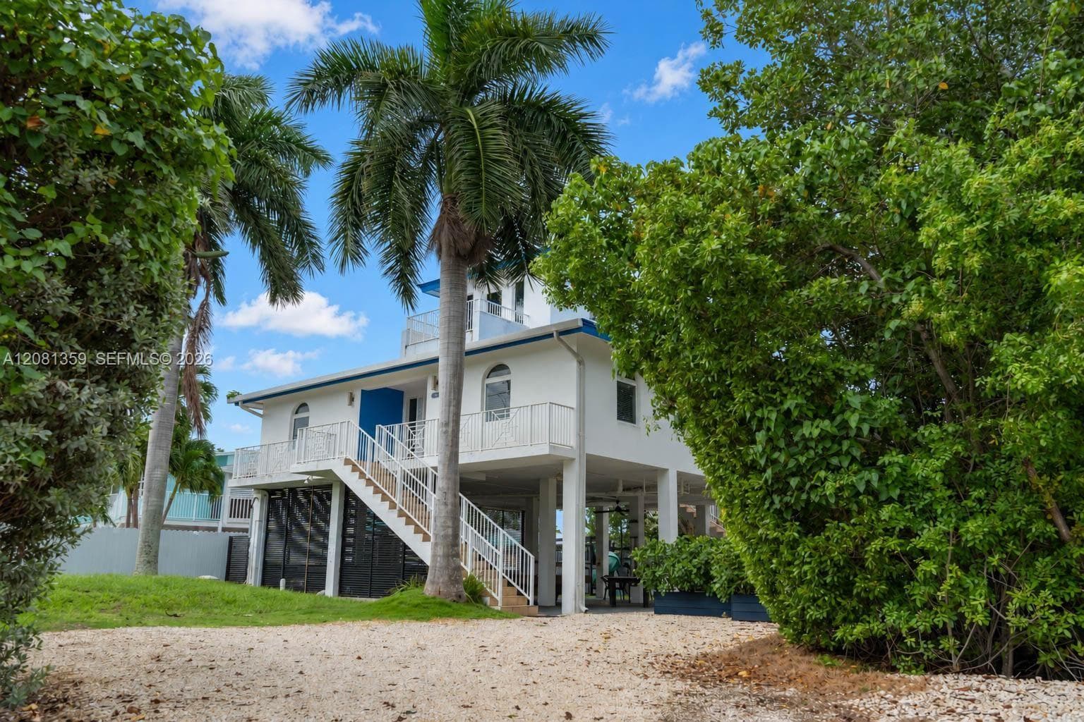 Stilt-construction residence featuring a white exterior, elevated wrap-around balcony with metal railings, external staircase, and ground-level covered patio area