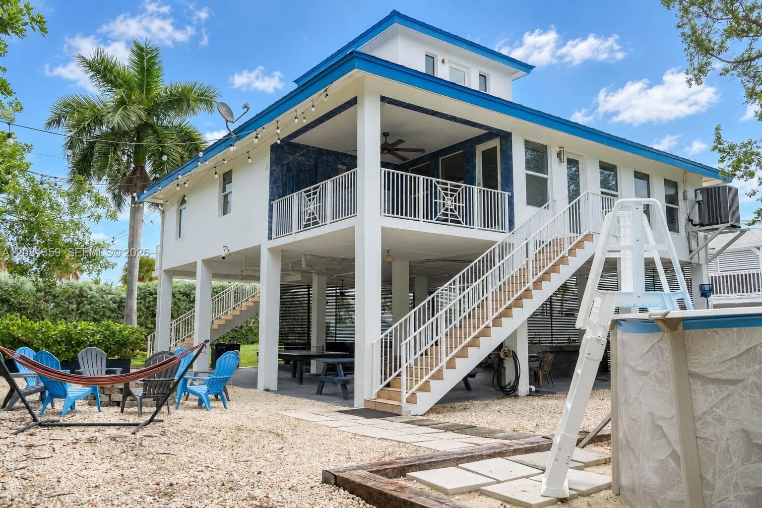 Elevated structure featuring a covered upper-level balcony with white metal railings, a paved ground-level patio area, exterior staircase with metal railings, and stone-ground landscaping