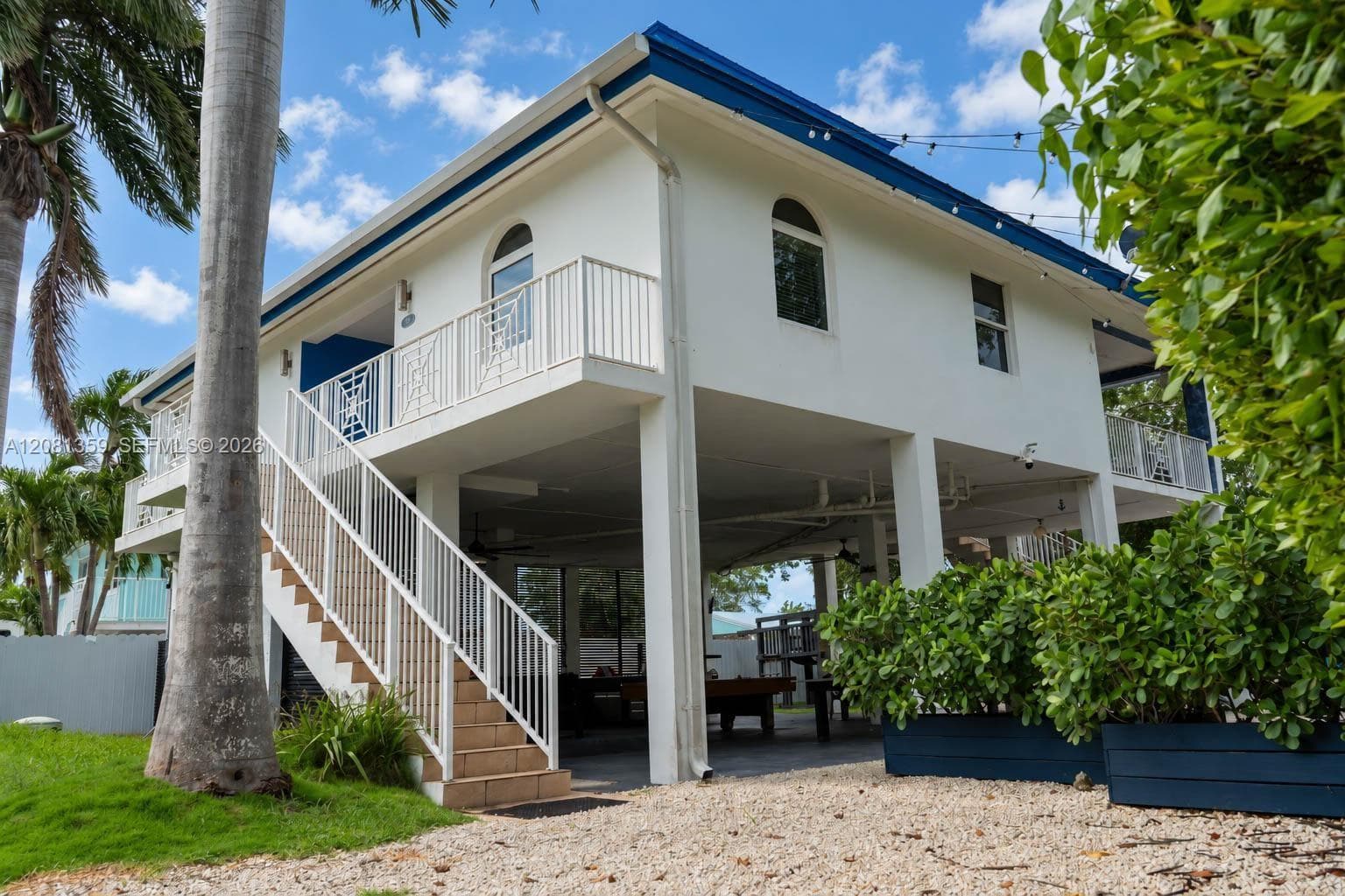 Stilted exterior featuring white stucco siding, blue roof accents, arched windows, a metal staircase with white railings, and a covered ground-level patio area