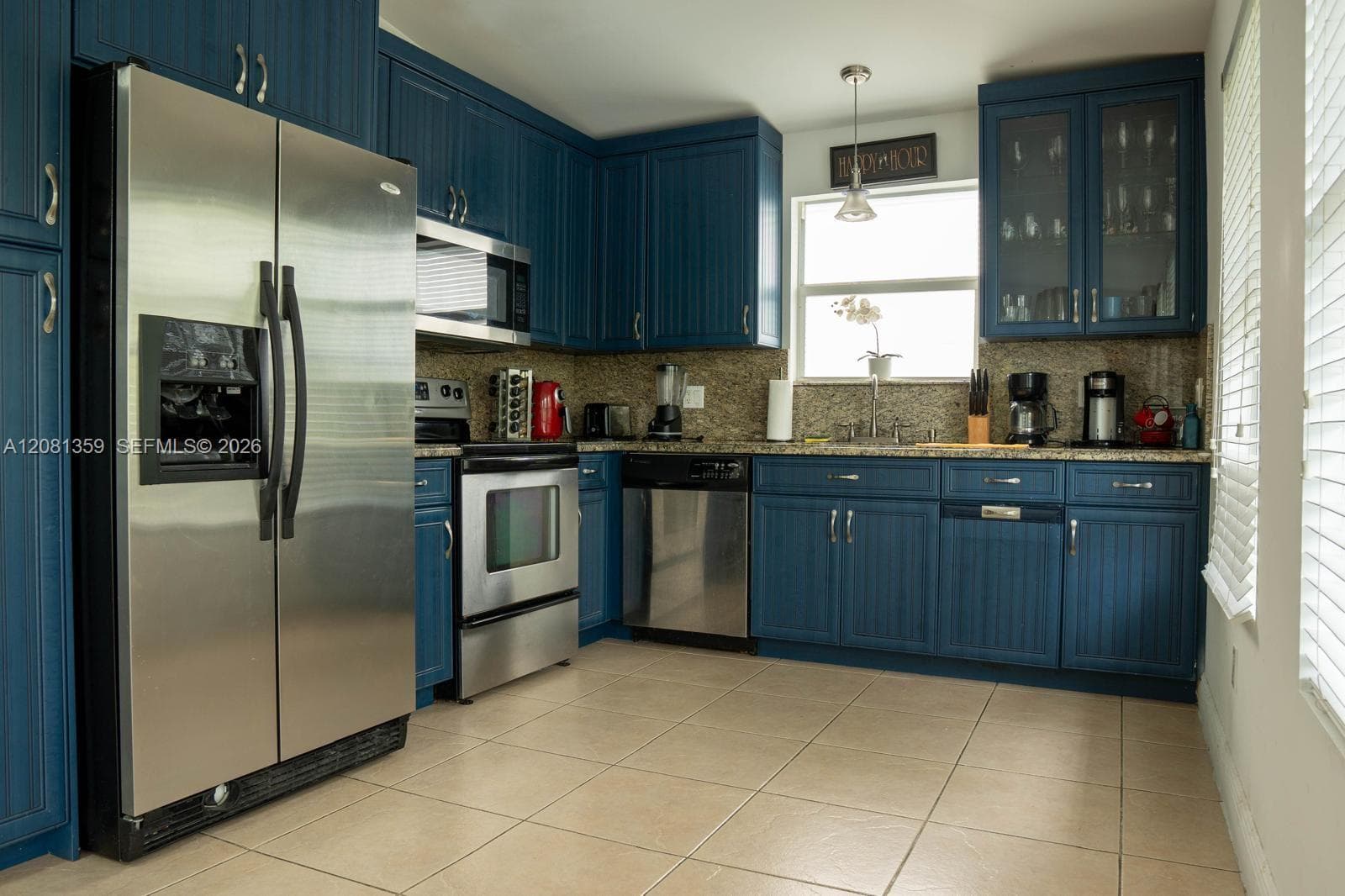 Kitchen featuring blue beadboard-style cabinetry, stainless steel appliances, stone-look countertops, glass-front upper cabinet displays, and ceramic tile flooring