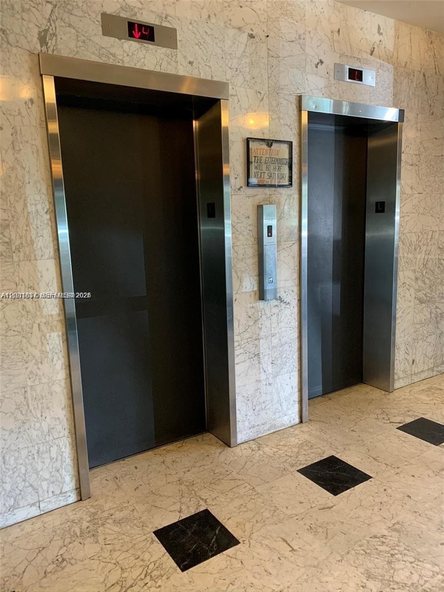Dual stainless steel elevator doors framed by polished stone wall cladding and stone tile flooring with decorative square inlays