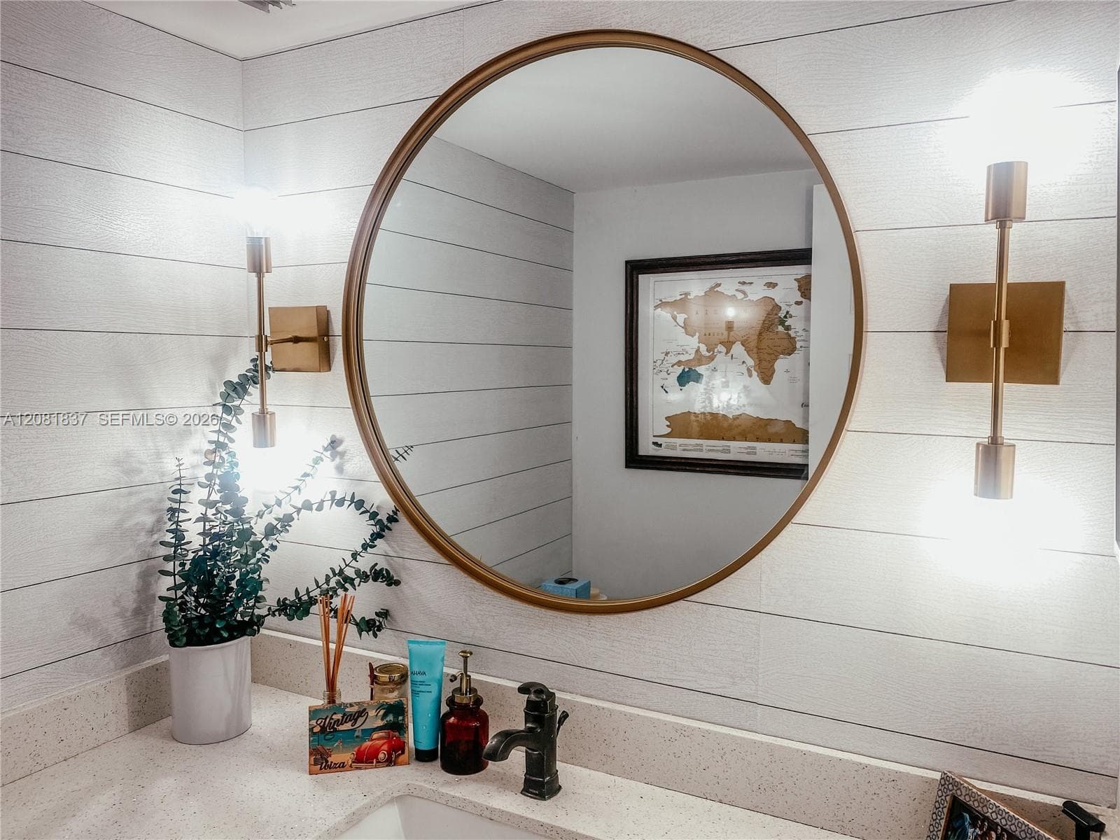 Round mirror with a metallic frame, horizontal plank wall cladding, integrated stone-surface countertop, and dual vertical wall-mounted fixtures with metallic finishes