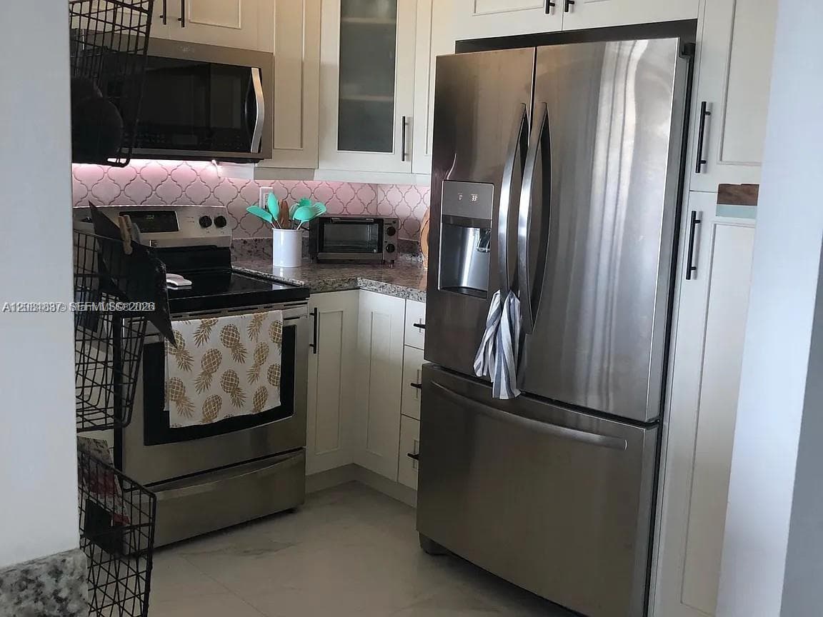 Kitchen featuring white shaker-style cabinetry, stainless steel appliances, speckled stone-finish countertops, patterned tile backsplash, and under-cabinet lighting