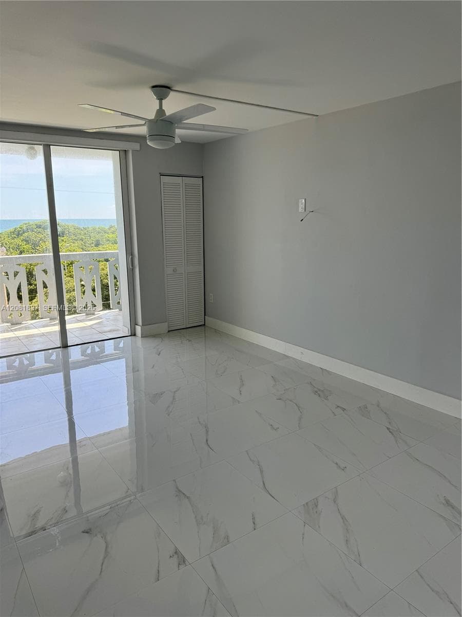 Polished large-format tile flooring with a marble-finish, white louvered closet door, ceiling fan with integrated lighting, and glass sliding doors opening to a balcony with an ocean backdrop