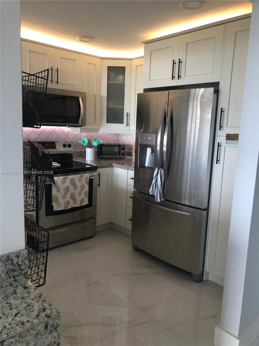 White cabinetry featuring glass-panel accents, stainless steel appliances, patterned tile backsplash, polished stone-finish flooring, and recessed ceiling lighting