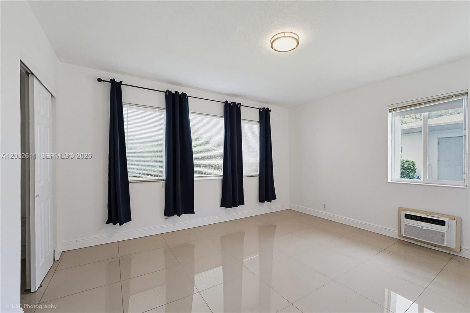 Neutral interior space featuring polished tile flooring, triple window configuration with a rod-mounted drapery system, an integrated wall-mounted climate control unit, and a flush-mount circular ceiling fixture