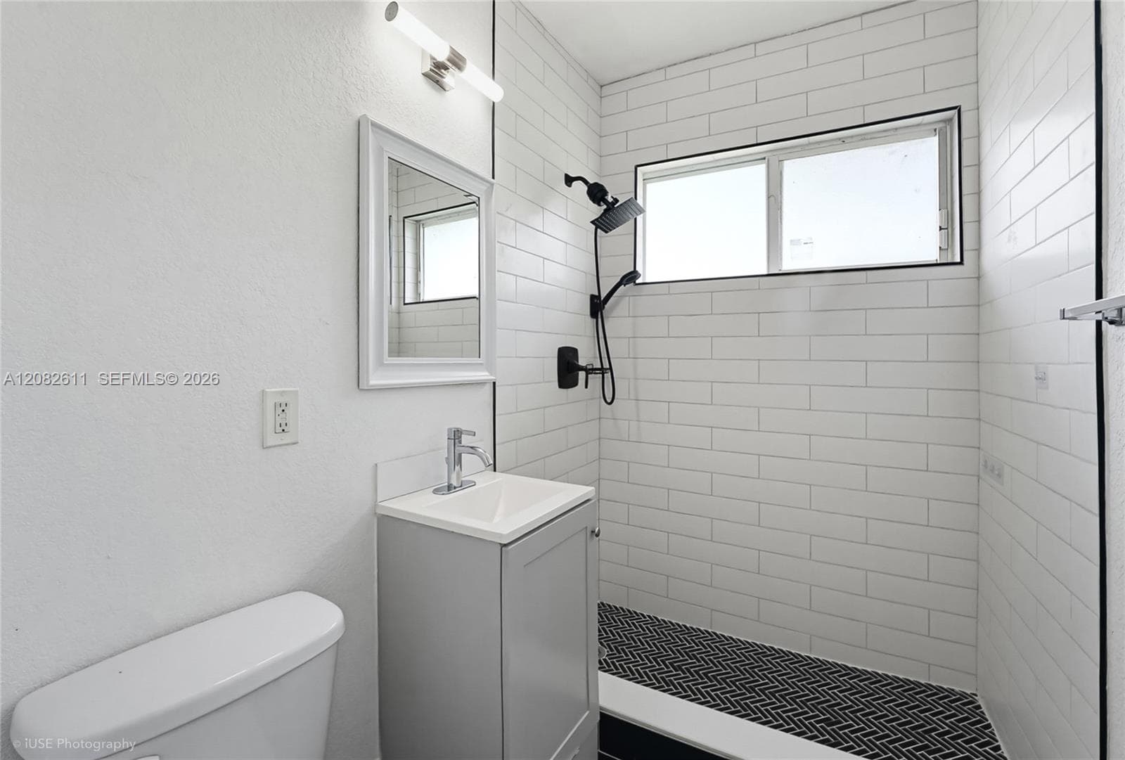 Bathroom featuring a grey vanity with a rectangular sink, white subway tile shower surround, matte black shower fixtures, and patterned tile flooring