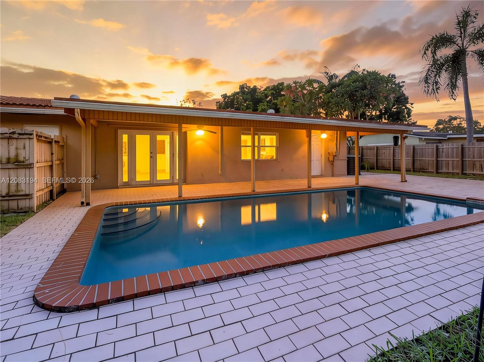 In-ground swimming pool with a brick-trimmed border, covered patio, light-colored paver decking, and wood-style privacy fencing