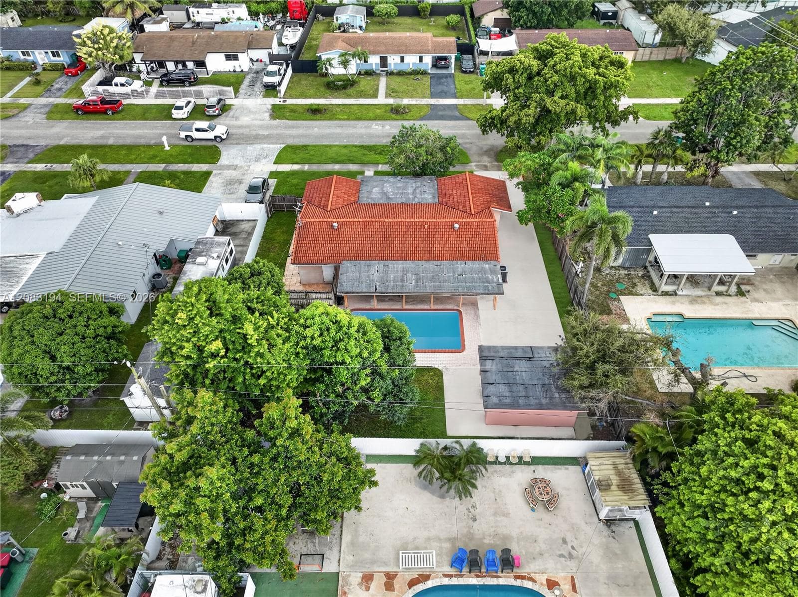 Aerial perspective of a property featuring a red tile roof, an in-ground swimming pool, a concrete patio, mature trees, and perimeter fencing