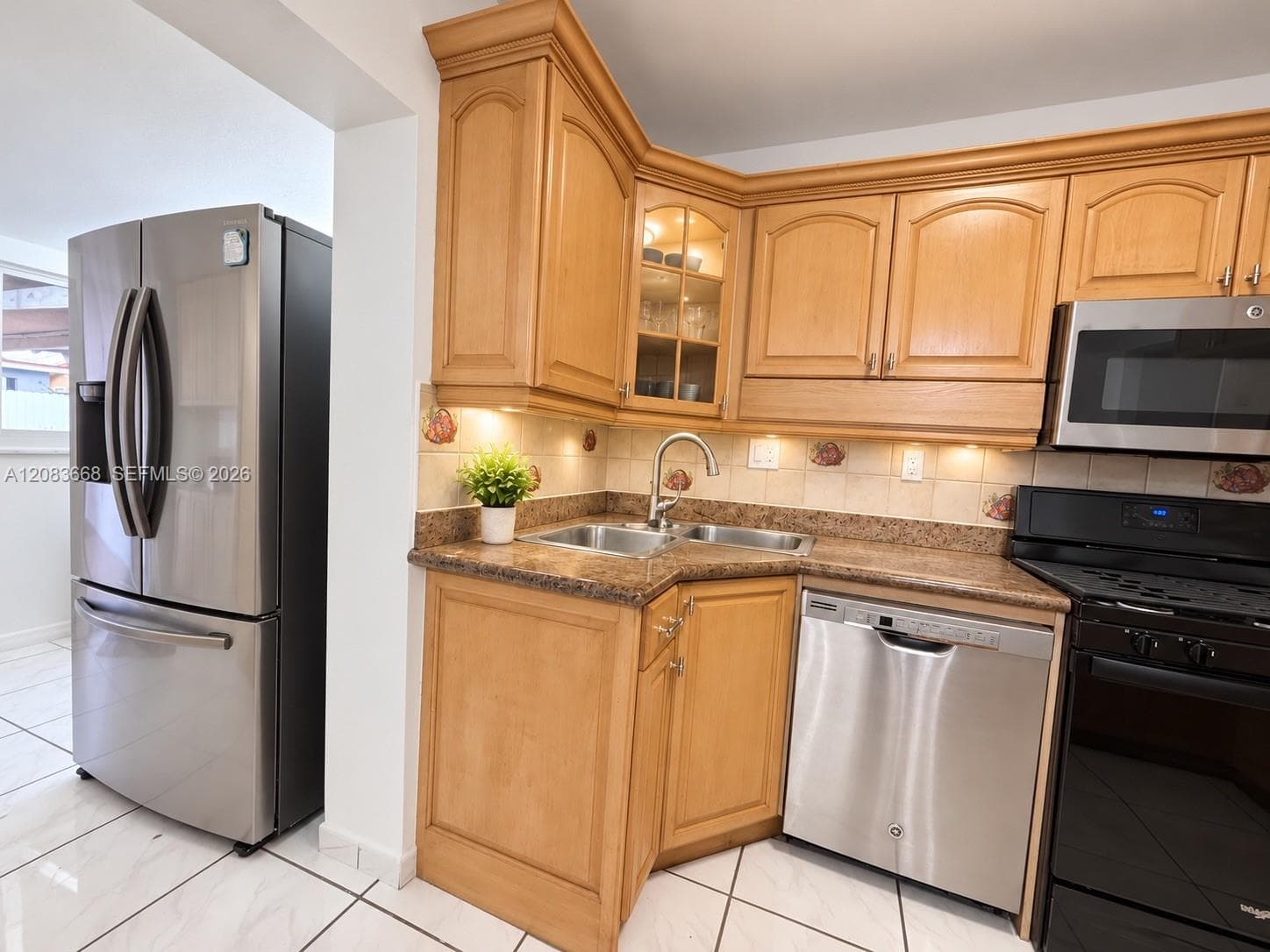 Wood-finish cabinetry with crown molding, stainless steel appliances, corner sink with gooseneck faucet, light-toned backsplash, and tile flooring