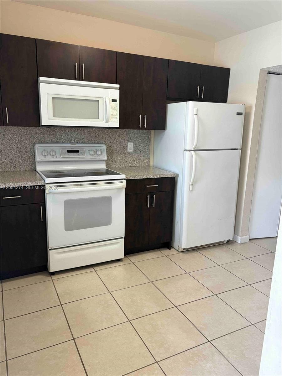 Dark wood-finish cabinetry, light-toned tiled flooring, white built-in microwave, white electric range, and white refrigerator