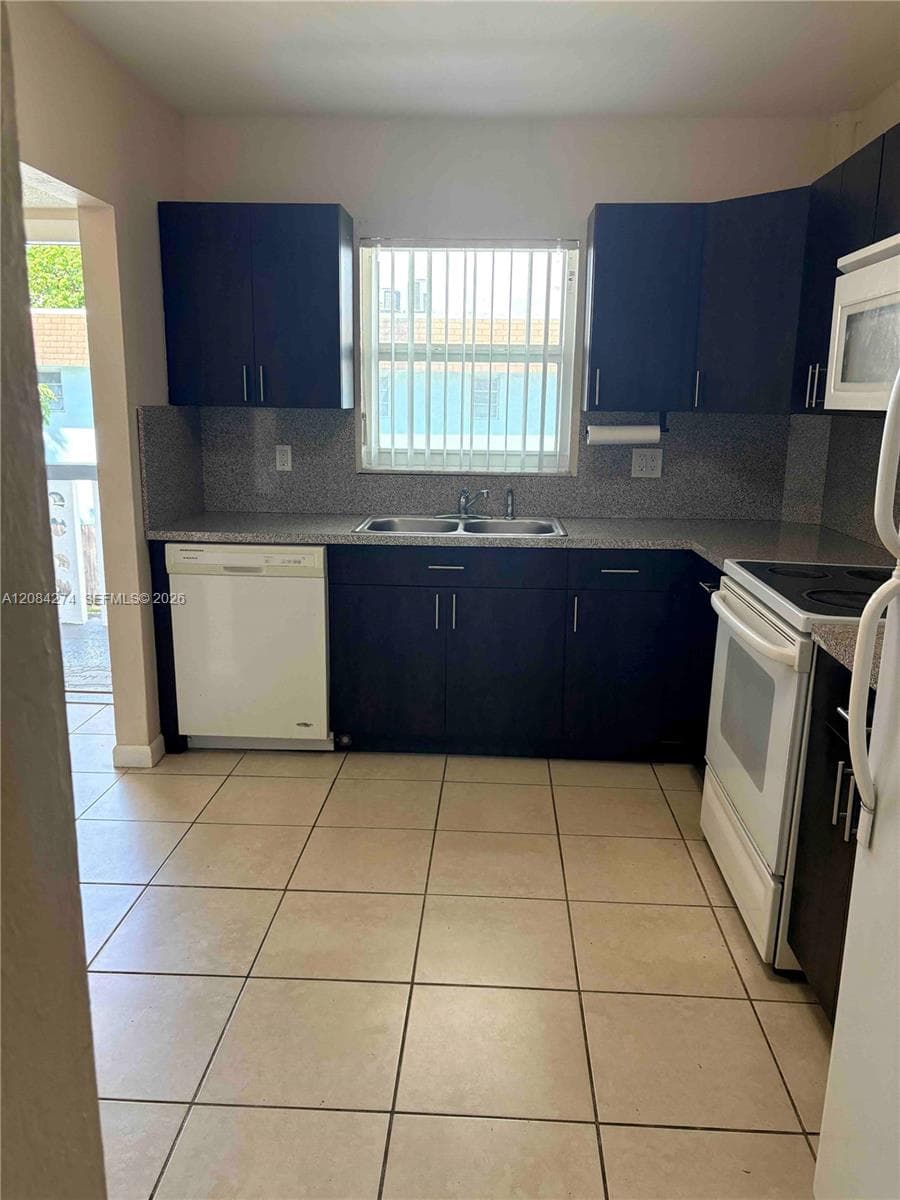 Ceramic tile flooring, dark wood-finish cabinetry, granite-look countertops, stainless steel double-basin sink, and a white dishwasher