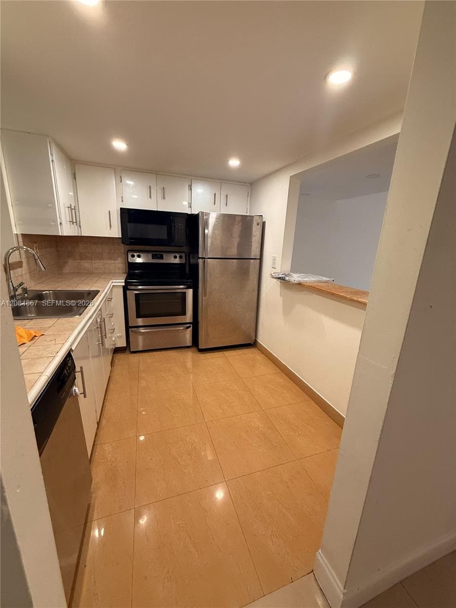 Kitchen featuring stainless steel appliances, white cabinetry, tile countertops, tile flooring, and a service pass-through window with a ledge