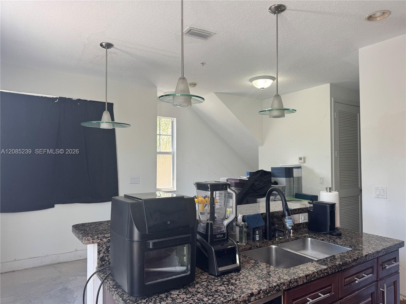 Kitchen island featuring stone countertops, a double-basin stainless steel sink with a high-arc faucet, dark wood-finish cabinetry, and three pendant lights with glass accents