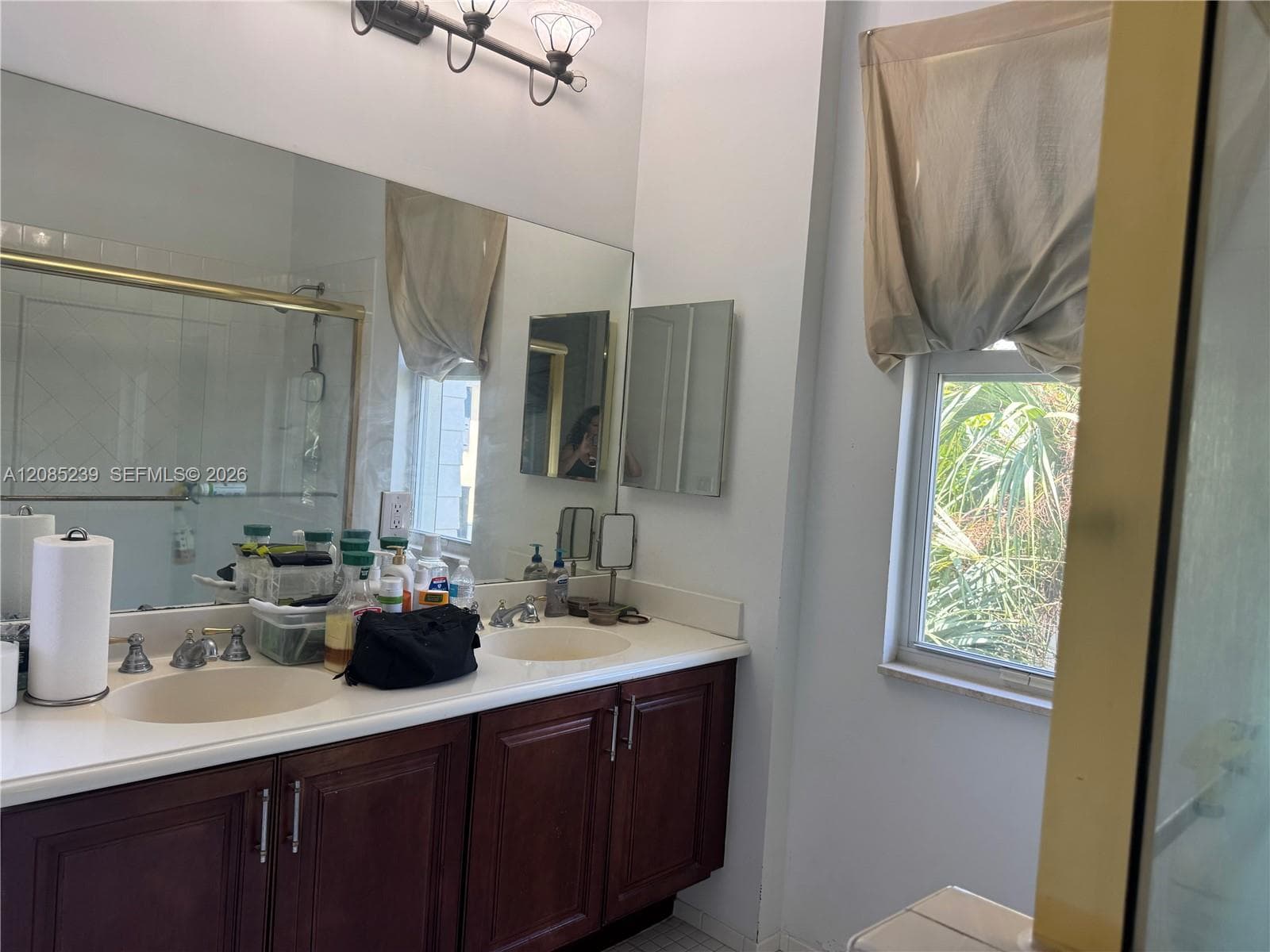 Double vanity featuring wood-finish cabinetry, solid-surface countertop with integrated dual sinks, large wall-mounted mirror, and three-bulb vanity lighting fixture