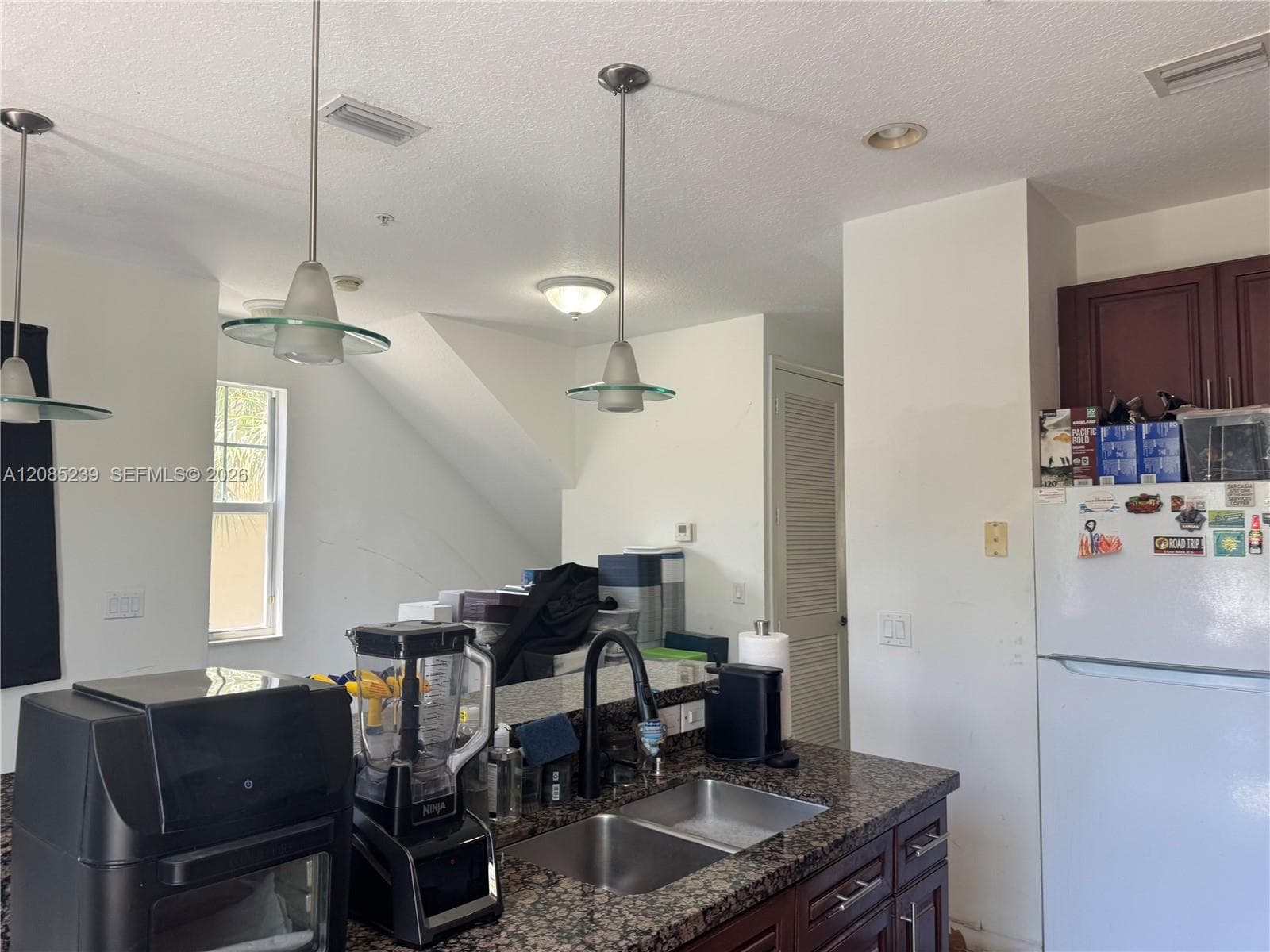 Kitchen featuring stone countertops, a dual-basin stainless steel sink with a high-arch pull-down faucet, dark wood-finish cabinetry, and glass-accented pendant lighting