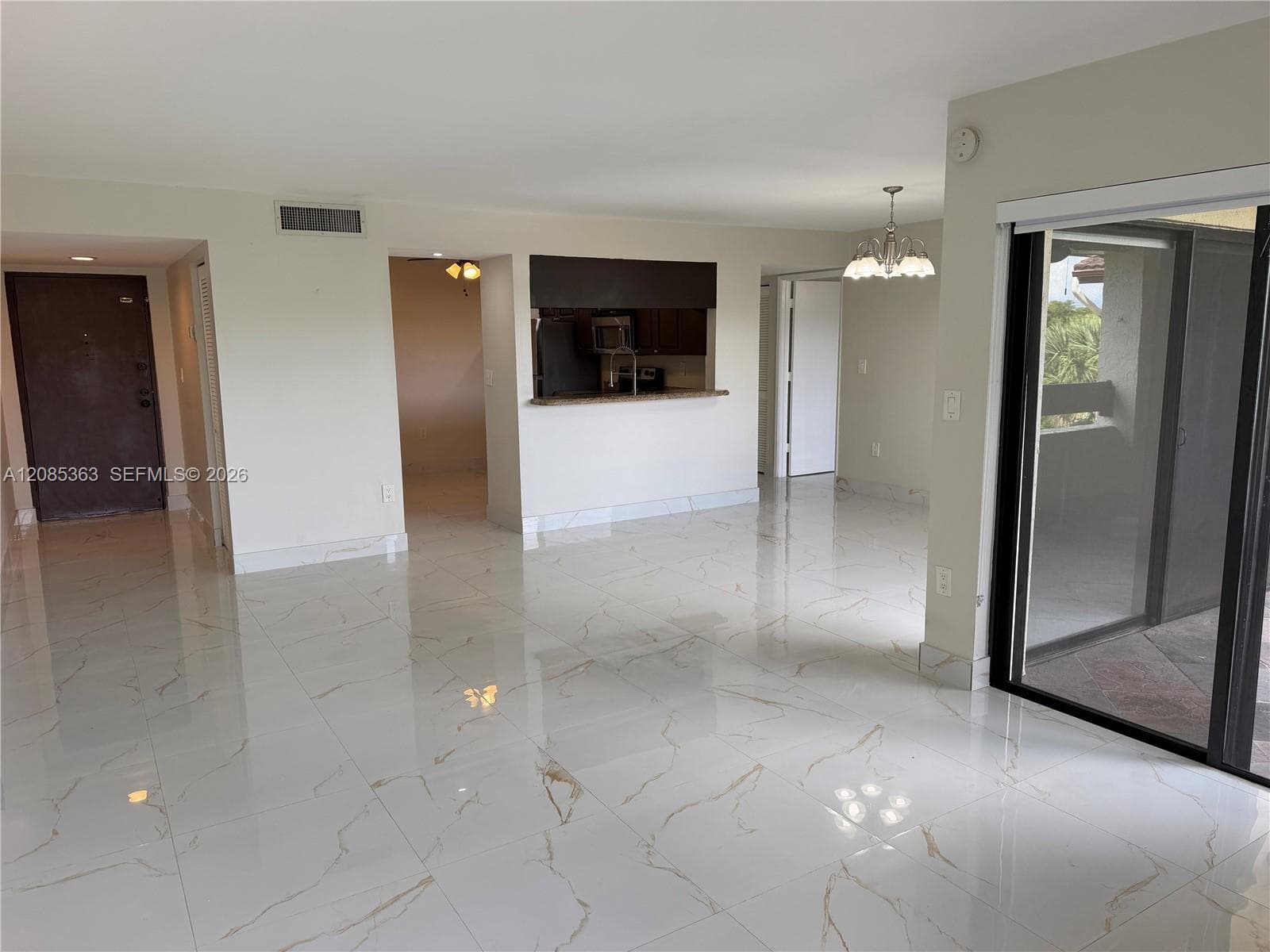 Open interior space featuring polished large-format porcelain tile flooring with veining, pass-through kitchen opening, built-in chandelier, and sliding glass door with dark trim