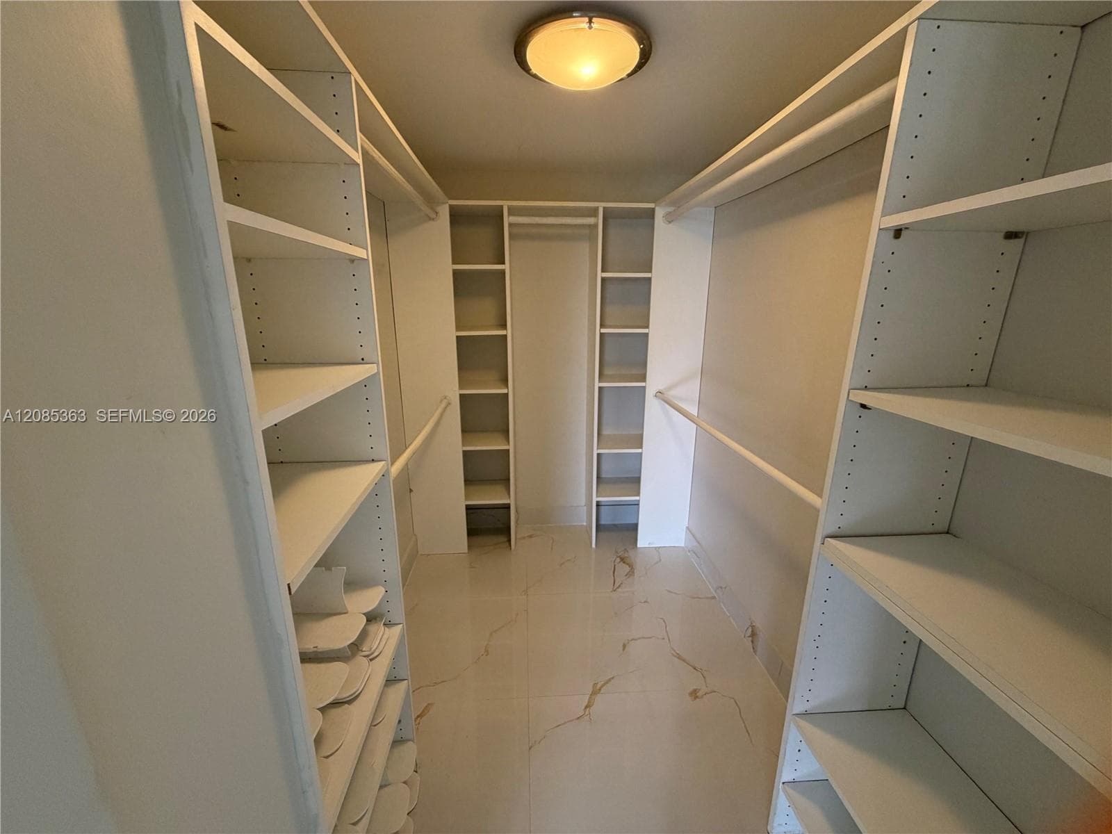 Walk-in closet featuring white built-in shelving, integrated hanging rods, flush-mount lighting, and large-format tile flooring