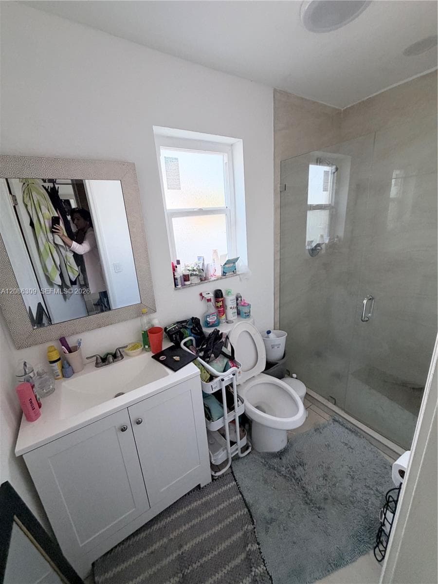 Bathroom featuring a white vanity cabinet with a composite countertop and integrated basin, a walk-in shower with a glass panel enclosure, and neutral wall tiling
