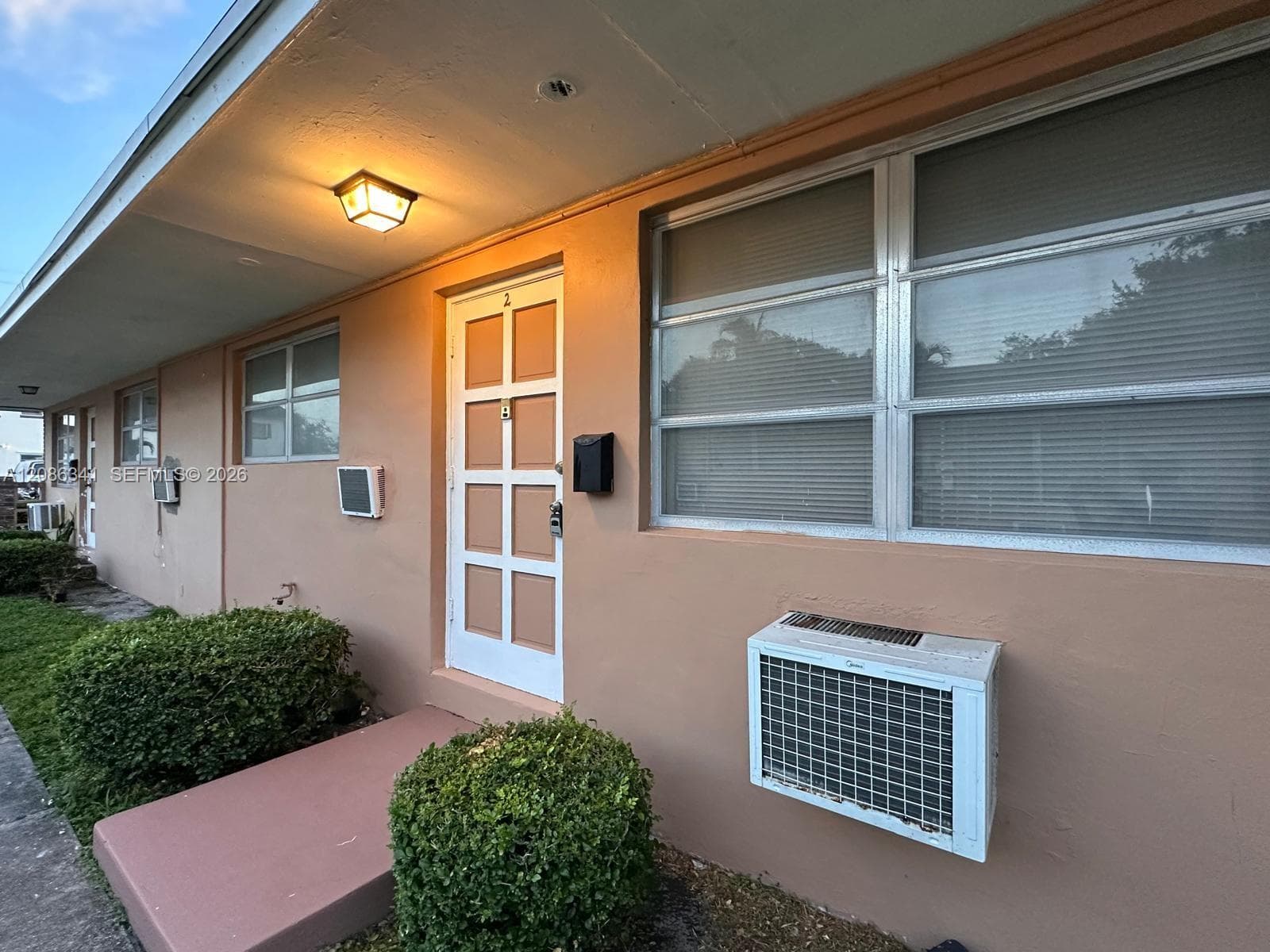 Exterior entry featuring a multi-pane glass door, horizontal sliding windows, a wall-mounted light fixture, and an integrated window air conditioning unit