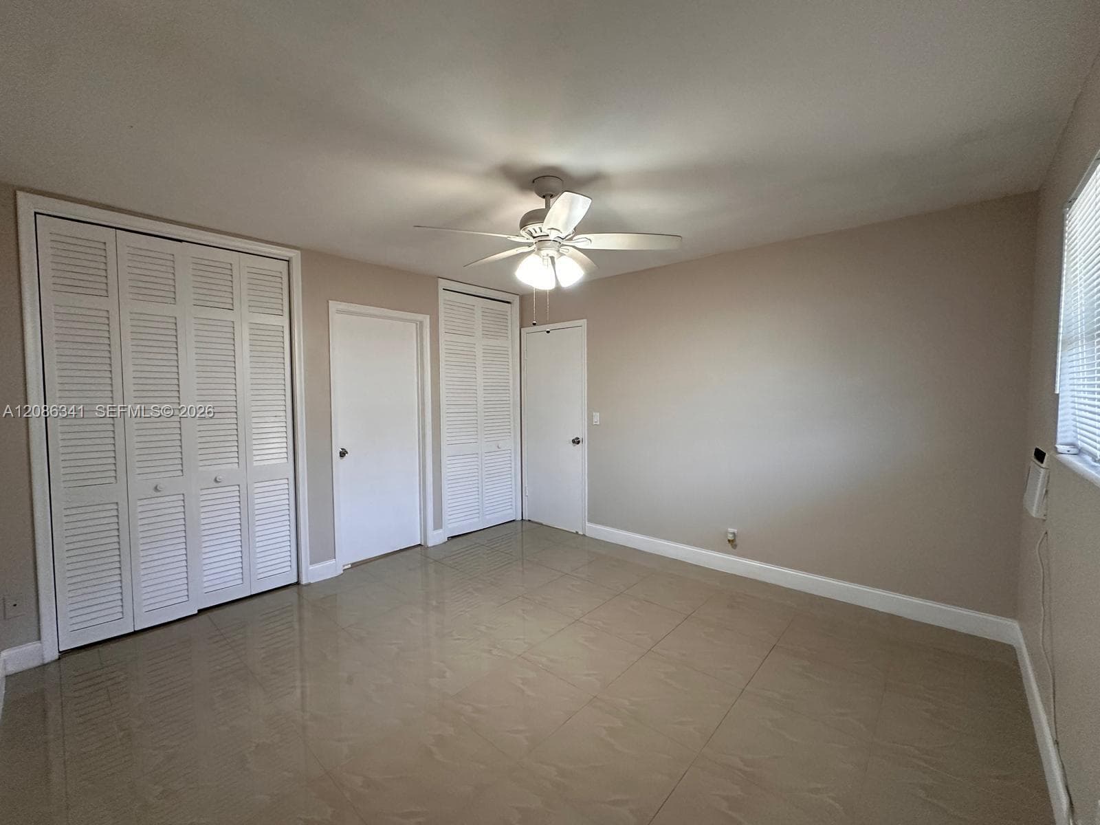 Large-format tile flooring, dual white louvered-door closets, single-panel interior doors, and a ceiling fan with integrated light fixture