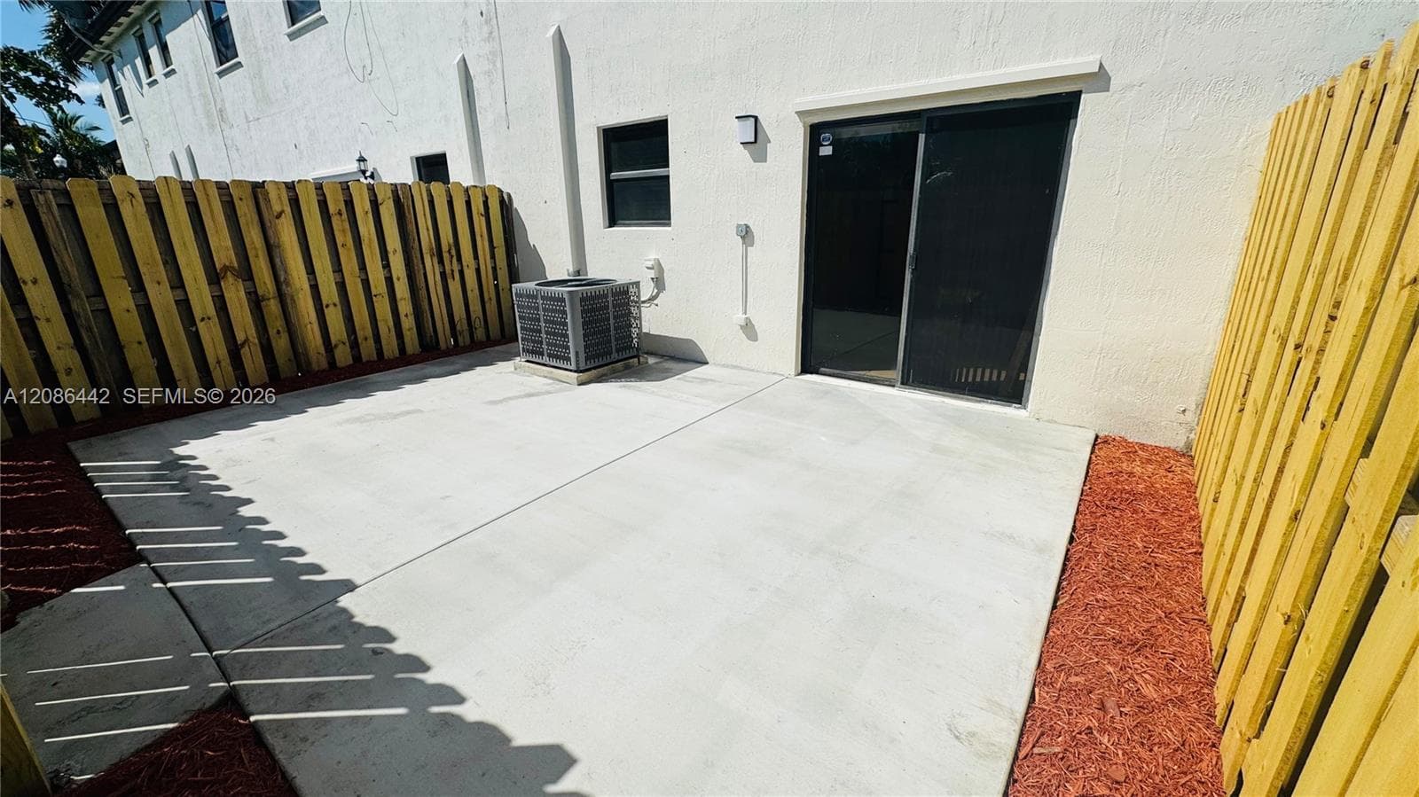 Private concrete patio featuring tall wood privacy fencing and red mulch borders, finished with a sliding glass door entry