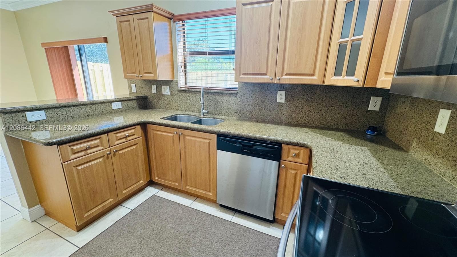 Kitchen featuring medium-tone wood cabinetry, stone-surface countertops, dual-basin stainless steel sink, tile flooring, and integrated stainless steel appliances