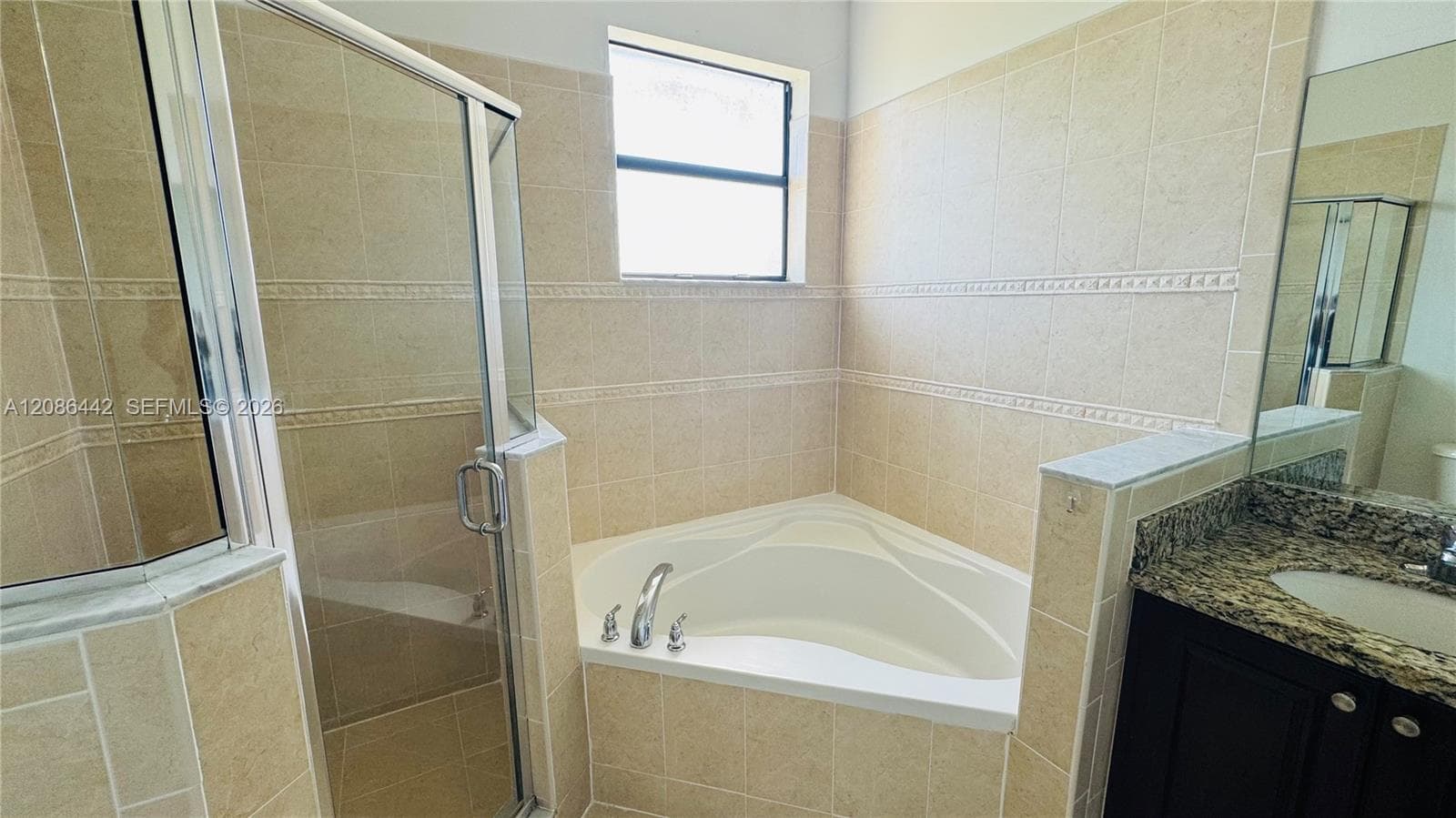 Corner soaking tub with tiled surround, glass-enclosed shower, dark wood-finish vanity, and stone-surface countertop