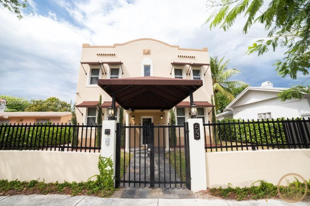 Stucco exterior featuring a arched roofline, covered entryway canopy, decorative window awnings, and a gated perimeter wall with stone-finish walkway