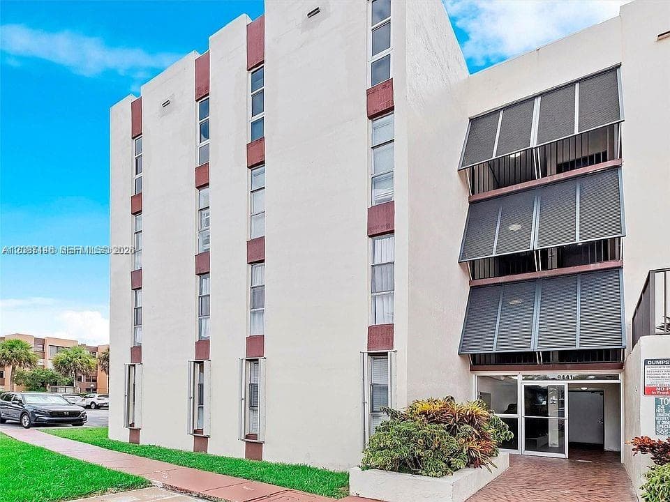 Mid-rise exterior featuring a textured stucco facade, vertical window alignments, metal balcony awnings, and a covered ground-level entryway